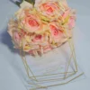 A transparent acrylic wedding invitation resting on a single, soft peach rose . The card is decorated with a bold gold geometric frame consisting of thin, intersecting lines that create a large diamond and angular border. The invitation text is printed in elegant gold foil.