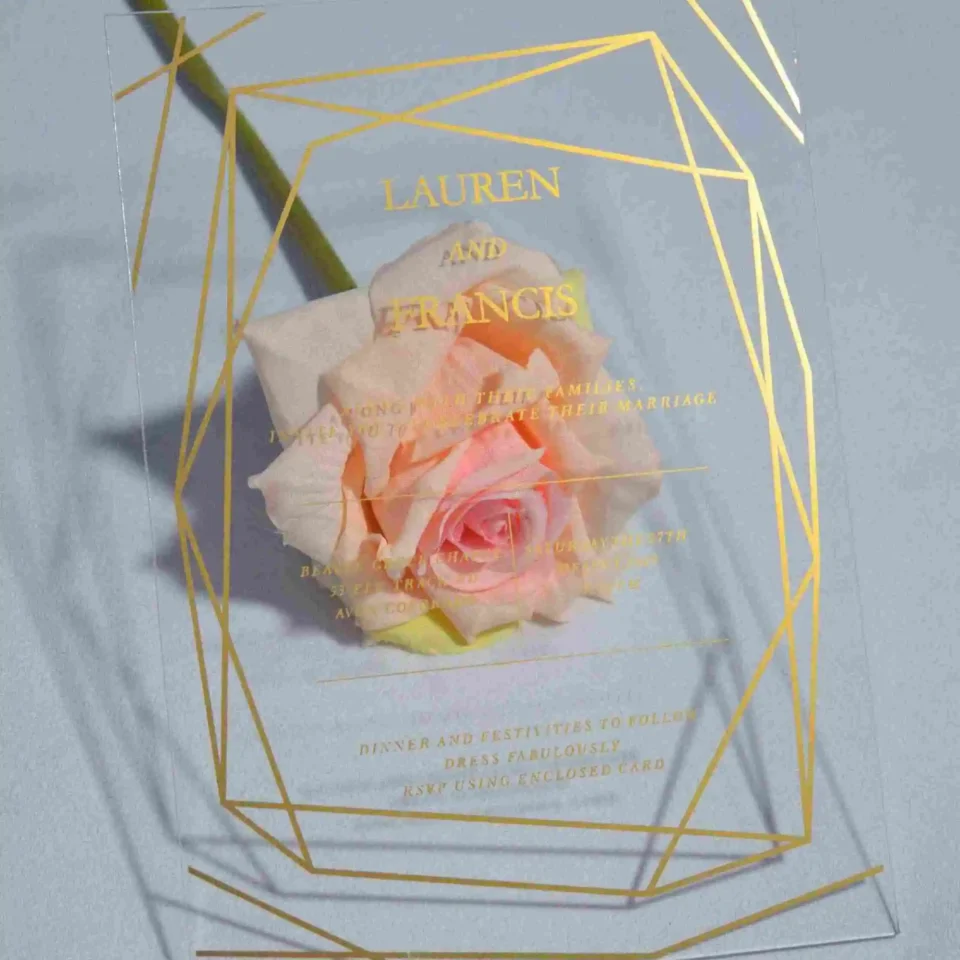 A transparent acrylic wedding invitation resting on a single, soft peach rose . The card is decorated with a bold gold geometric frame consisting of thin, intersecting lines that create a large diamond and angular border. The invitation text is printed in elegant gold foil.