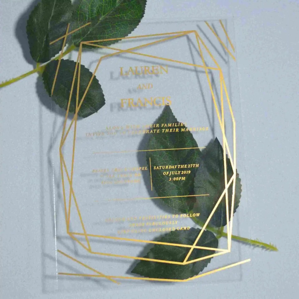 A transparent acrylic wedding invitation resting on a single, soft green leaves. The card is decorated with a bold gold geometric frame consisting of thin, intersecting lines that create a large diamond and angular border. The invitation text is printed in elegant gold foil.