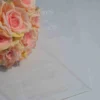 A transparent, rectangular acrylic wedding invitation is laid flat over several green leaves on a white surface. The invitation features clean, white serif text that appears to float above the organic textures of the foliage.