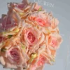A transparent acrylic wedding invitation is shown leaning against a large bouquet of soft peach and pink roses. The invitation features clean white printed text with a minimalist layout.