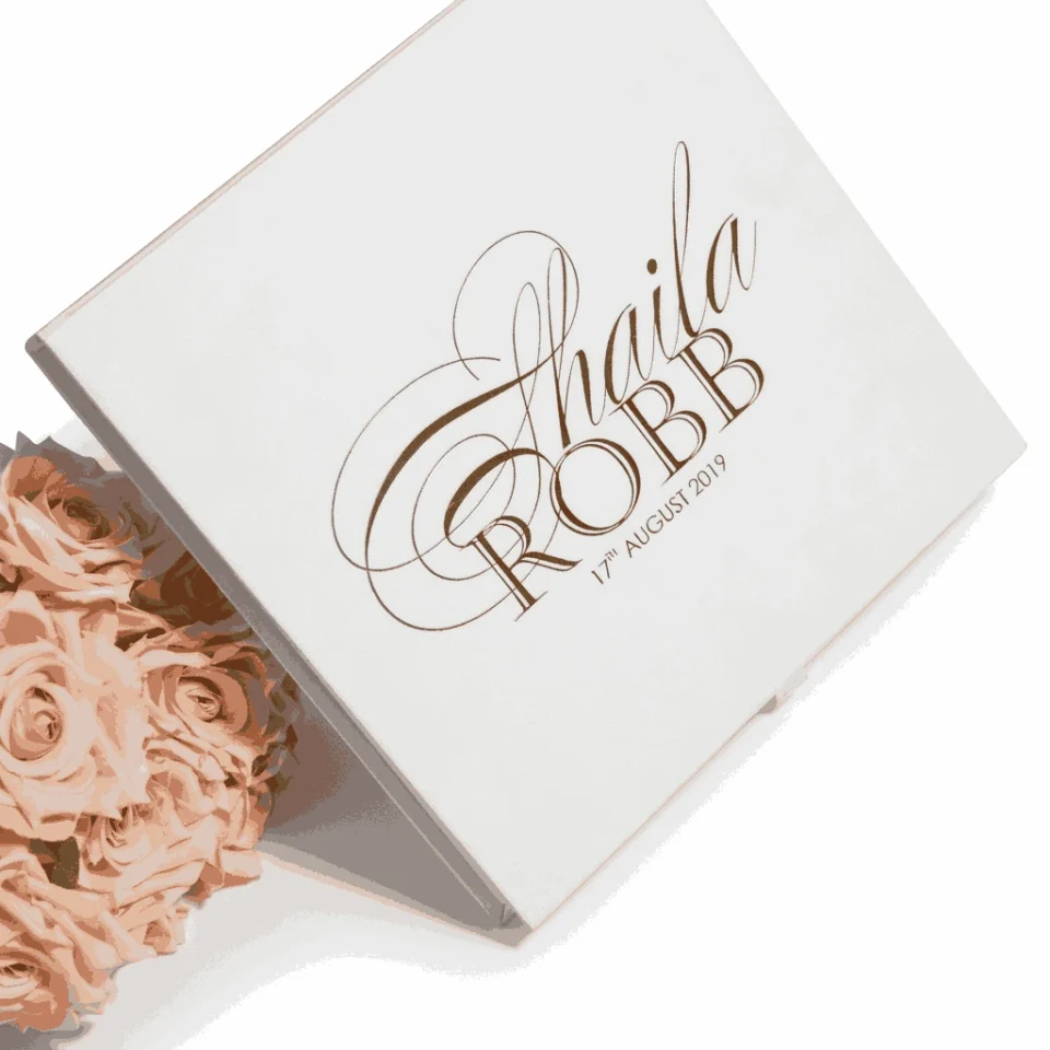 An open, square ivory silk invitation box featuring a multi-layered stationery suite inside. The main wedding invitation for Victoria Marie and Patric Johnson is nestled in the base, framed by an elegant black scrollwork border. The lid is propped open, revealing an interior pocket that holds an RSVP card and a smaller card with the names "Oliver and Emily" in script. The entire set is shown against a clean white background, emphasizing the luxurious fabric texture and crisp typography.