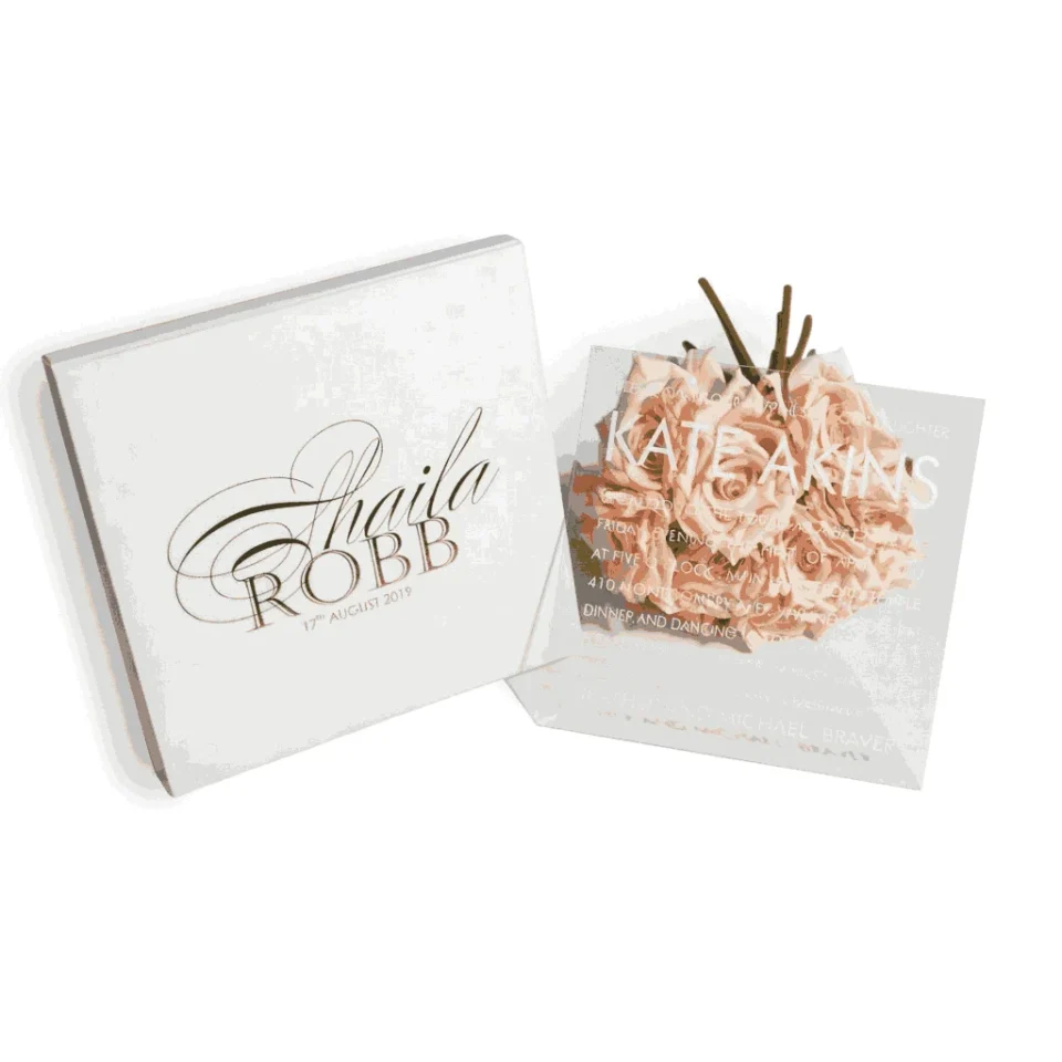 A high-angle shot of an open, square ivory silk invitation box. The lid is propped open, featuring an interior pocket that holds multiple stationery items, including an RSVP card and a small card with the names "Oliver and Emily." In the base of the box, a formal wedding invitation for Victoria Marie and Patric Johnson is displayed, decorated with ornate black scrollwork at the corners. The box is set on a white surface scattered with light-colored dried petals, and a stem of white gladiolus flowers lies to the right.