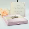 An open, light pink silk invitation box containing a celebration card for Arlyn Estrella Bac. The card features elegant silver scrollwork at the corners and black script typography. The box is shown against a clean white background, surrounded by white and soft peach-colored roses, with a small portion of a bronze Eiffel Tower figurine visible on the right.