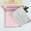 An open, light pink silk invitation box containing a celebration card for Arlyn Estrella Bac. The card features elegant silver scrollwork at the corners and black script typography. The box is shown against a clean white background, surrounded by white and soft peach-colored roses, with a small portion of a bronze Eiffel Tower figurine visible on the right.