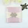 A top-down view of a square, dusty rose silk invitation folio resting on a white surface. An ivory satin ribbon is wrapped horizontally across the center, fastened with a large, ornate silver brooch encrusted with clear crystals and teardrop-shaped rhinestones. In the upper corners, two white artificial roses are partially visible, and a small bronze Eiffel Tower figurine sits to the right of the folio.