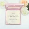 An open, light pink silk invitation box containing a celebration card for Arlyn Estrella Bac. The card features elegant silver scrollwork at the corners and black script typography. The box is shown against a clean white background, surrounded by white and soft peach-colored roses, with a small portion of a bronze Eiffel Tower figurine visible on the right.