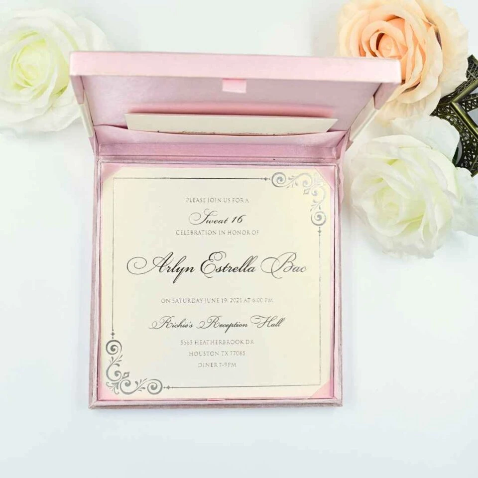 An open, light pink silk invitation box containing a celebration card for Arlyn Estrella Bac. The card features elegant silver scrollwork at the corners and black script typography. The box is shown against a clean white background, surrounded by white and soft peach-colored roses, with a small portion of a bronze Eiffel Tower figurine visible on the right.