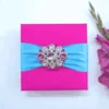 A vibrant, hot pink square invitation box featuring a wide, light blue satin ribbon wrapped horizontally across the center. The ribbon is fastened with a large, ornate silver brooch encrusted with pink crystals and rhinestones. The box is set against a clean white background, with a stem of bright pink gladiolus flowers lying vertically along the right side.