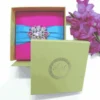A vibrant, hot pink square invitation box featuring a wide, light blue satin ribbon wrapped horizontally across the center. The ribbon is fastened with a large, ornate silver brooch encrusted with pink crystals and rhinestones. The box is set against a clean white background, with a stem of bright pink gladiolus flowers lying vertically along the right side.