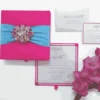 A top-down view of a square, blush pink silk invitation folio resting on a white surface. An light blue satin ribbon is wrapped horizontally across the center, fastened with a large, ornate silver brooch encrusted with clear crystals and sparkling rhinestones. In the upper corners, two white artificial roses are partially visible, and a small bronze Eiffel Tower figurine sits to the right of the folio.
