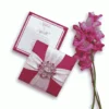 A high-angle shot featuring a square, deep magenta invitation box and its matching invitation card. The closed box is decorated with wide white satin ribbons crossing in the center, secured by a large, ornate silver brooch encrusted with clear and pink crystals. To the left, the invitation card for "Mathew and Michelle" is displayed, featuring elegant pink script and a magenta border. A stem of vibrant pink gladiolus flowers lies vertically along the right side of the arrangement against the white background.
