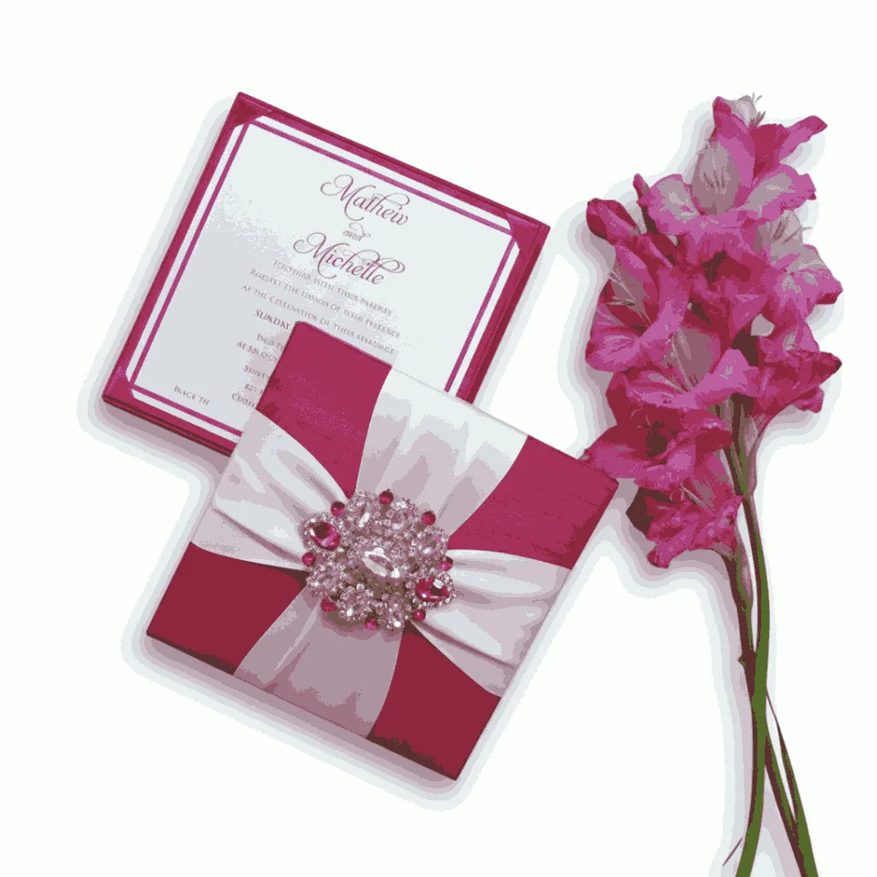 A high-angle shot featuring a square, deep magenta invitation box and its matching invitation card. The closed box is decorated with wide white satin ribbons crossing in the center, secured by a large, ornate silver brooch encrusted with clear and pink crystals. To the left, the invitation card for "Mathew and Michelle" is displayed, featuring elegant pink script and a magenta border. A stem of vibrant pink gladiolus flowers lies vertically along the right side of the arrangement against the white background.