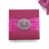 A high-angle shot of a square, textured magenta silk invitation box. A vibrant pink satin ribbon is wrapped horizontally across the center, cinched by a large, circular silver brooch featuring a radiating pattern of clear, marquise-cut crystals. The arrangement is set against a clean white background, with a cluster of bright pink gladiolus flowers partially visible in the top right corner.
