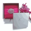 A high-angle shot of a square, textured magenta silk invitation box. A vibrant pink satin ribbon is wrapped horizontally across the center, cinched by a large, circular silver brooch featuring a radiating pattern of clear, marquise-cut crystals. The arrangement is set against a clean white background, with a cluster of bright pink gladiolus flowers partially visible in the top right corner.