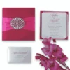 A flat-lay arrangement featuring a closed magenta silk invitation box decorated with a pink ribbon and a large circular crystal brooch. Surrounding the box are a matching wedding invitation, an RSVP card, and an envelope for "Mathew and Michelle." A stem of bright pink gladiolus flowers lies in the bottom left corner against a white background.