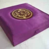 A low-angle shot of a square, vibrant purple silk invitation box resting on a marbled white surface. The padded lid features a central circular acrylic plaque in deep burgundy with a gold-foil border. The plaque is decorated with an elegant gold monogram "R & L" intertwined with delicate floral illustrations.