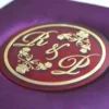A low-angle shot of a square, vibrant purple silk invitation box resting on a marbled white surface. The padded lid features a central circular acrylic plaque in deep burgundy with a gold-foil border. The plaque is decorated with an elegant gold monogram "R & L" intertwined with delicate floral illustrations.
