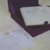 An open, square purple silk invitation box resting on a white marbled surface. Nested inside the box is a formal white wedding invitation for Thomas and Wallis, featuring a gold-scrolled border and a central gold monogram. In the foreground, a matching white RSVP card is displayed, decorated with gold floral scrollwork and an "RSVP" heading in elegant script. Another card with the names Victoria and Patric is partially visible on the left.