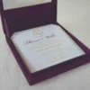 An open, square purple silk invitation box resting on a white marbled surface. Nested inside the box is a formal white wedding invitation for Thomas and Wallis, featuring a gold-scrolled border and a central gold monogram. In the foreground, a matching white RSVP card is displayed, decorated with gold floral scrollwork and an "RSVP" heading in elegant script. Another card with the names Victoria and Patric is partially visible on the left.