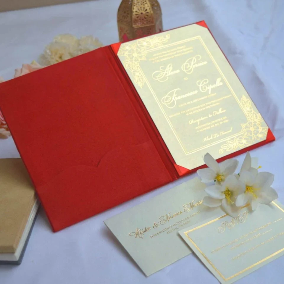 An open red velvet wedding invitation folio showcased on a white surface. The interior features a scalloped pocket on the left and a stunning gold metallic invitation card on the right, held in place by red satin corner ribbons. The gold card is detailed with ornate floral engravings and elegant script for the names.
