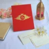 A luxurious, vibrant red velvet wedding invitation box or folio resting on a white fabric surface. The cover features an intricate gold foil crest in the center, containing a monogrammed topped with a royal crown and surrounded by ornate filigree.