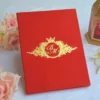 A luxurious, vibrant red velvet wedding invitation box or folio resting on a white fabric surface. The cover features an intricate gold foil crest in the center, containing a monogrammed topped with a royal crown and surrounded by ornate filigree.
