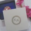 A top-down view of a square, royal blue silk invitation box resting on a textured white surface. A matching royal blue satin ribbon is wrapped horizontally across the center, cinched by a large, ornate gold brooch encrusted with clear and pink crystals. To the right of the box, a stem of vibrant pink gladiolus flowers adds a soft touch to the composition.