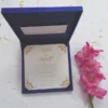A top-down view of a square, royal blue silk invitation box set against a white background. A matching blue satin ribbon is wrapped horizontally across the center, cinched by a large, ornate silver brooch encrusted with clear and pink crystals. A stem of vibrant pink gladiolus flowers lies vertically along the right side of the box.