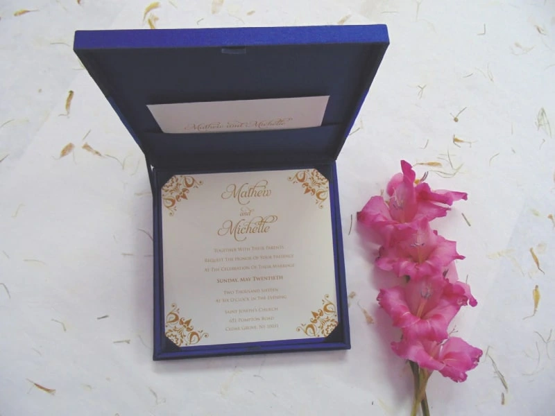 A top-down view of a square, royal blue silk invitation box set against a white background. A matching blue satin ribbon is wrapped horizontally across the center, cinched by a large, ornate silver brooch encrusted with clear and pink crystals. A stem of vibrant pink gladiolus flowers lies vertically along the right side of the box.