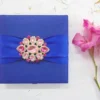 A top-down view of a square, royal blue silk invitation box resting on a textured white surface. A matching royal blue satin ribbon is wrapped horizontally across the center, cinched by a large, ornate gold brooch encrusted with clear and pink crystals. To the right of the box, a stem of vibrant pink gladiolus flowers adds a soft touch to the composition.