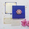 A top-down view of a square, royal blue silk invitation box set against a white background. A matching blue satin ribbon is wrapped horizontally across the center, cinched by a large, ornate silver brooch encrusted with clear and pink crystals. A stem of vibrant pink gladiolus flowers lies vertically along the right side of the box.