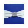 A top-down view of a square, navy blue silk invitation folio resting on a white surface. An ivory satin ribbon is wrapped horizontally across the center, fastened with a large, ornate silver brooch encrusted with clear crystals and teardrop-shaped rhinestones. In the upper corners, two white artificial roses are partially visible, and a small bronze Eiffel Tower figurine sits to the right of the folio.