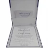 A flat-lay arrangement of royal blue wedding stationery on a textured white surface. It features a closed blue silk box with a matching ribbon and an ornate gold and pink crystal brooch. Surrounding the box are a formal white invitation with gold accents, a matching RSVP card, and a white envelope. A stem of pink flowers lies in the bottom right corner.