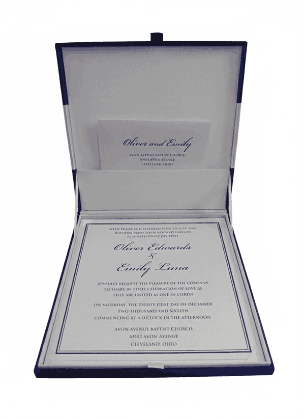 A flat-lay arrangement of royal blue wedding stationery on a textured white surface. It features a closed blue silk box with a matching ribbon and an ornate gold and pink crystal brooch. Surrounding the box are a formal white invitation with gold accents, a matching RSVP card, and a white envelope. A stem of pink flowers lies in the bottom right corner.