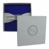 A top-down view of a square, navy blue silk invitation folio resting on a white surface. An ivory satin ribbon is wrapped horizontally across the center, fastened with a large, ornate silver brooch encrusted with clear crystals and teardrop-shaped rhinestones. In the upper corners, two white artificial roses are partially visible, and a small bronze Eiffel Tower figurine sits to the right of the folio.