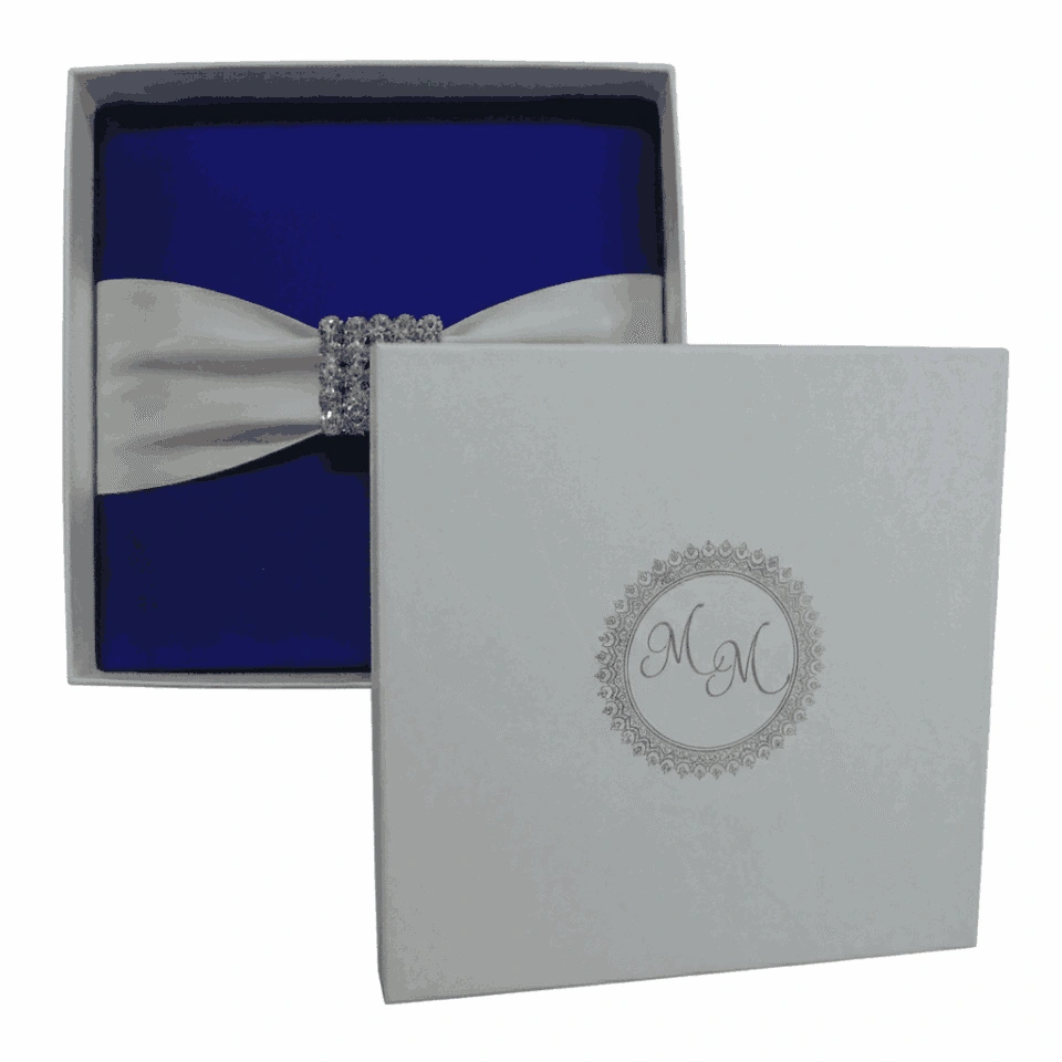 A top-down view of a square, navy blue silk invitation folio resting on a white surface. An ivory satin ribbon is wrapped horizontally across the center, fastened with a large, ornate silver brooch encrusted with clear crystals and teardrop-shaped rhinestones. In the upper corners, two white artificial roses are partially visible, and a small bronze Eiffel Tower figurine sits to the right of the folio.