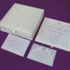 Here is the ALT text for the final image in your collection: **DSC_0293_165_11zon-960x804.jpg** An open, square ivory-colored wedding invitation box resting on a textured purple fabric surface. The interior lid features a horizontal pocket containing a small card for "Gabriella and Andre." The base of the box holds the main wedding invitation for "Gabriella Milagros Levin and Andre Leon Keavera," printed in elegant script on a white card with delicate corner flourishes.