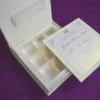 Here is the ALT text for the final image in your collection: **DSC_0294_161_11zon-960x706.jpg** An open, ivory-colored invitation box set against a textured purple background. The interior of the lid features a horizontal pocket holding a matching name card. The base of the box contains a white wedding invitation for "Gabriella Milagros Levin and Andre Leon Keavera," which is tilted to reveal a recessed grid with six empty compartments underneath, designed for holding favors or chocolates.
