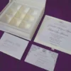 Here is the ALT text for the final image in your collection: **DSC_0294_161_11zon-960x706.jpg** An open, ivory-colored invitation box set against a textured purple background. The interior of the lid features a horizontal pocket holding a matching name card. The base of the box contains a white wedding invitation for "Gabriella Milagros Levin and Andre Leon Keavera," which is tilted to reveal a recessed grid with six empty compartments underneath, designed for holding favors or chocolates.