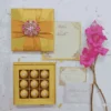 A top-down flat-lay arrangement of a gold-themed wedding stationery and gift set on a white background. The set features a closed square gold silk box decorated with a cross-shaped gold ribbon and a large, ornate crystal brooch. Next to it, the open base of a matching box reveals a 16-compartment tray filled with gold-foil wrapped chocolate truffles. Below the boxes are a formal white invitation for "Mathew and Michelle" with gold accents, a matching RSVP card, and a white envelope. A stem of bright pink gladiolus flowers lies along the right side of the frame.