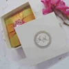A top-down photograph of a closed square gold satin invitation box on a mottled white background with loose petals. The lid is wrapped with a wide, cross-shaped gold satin ribbon, cinched at the intersection by a large, ornate gold brooch heavily encrusted with sparkling clear and pink crystals in a starburst pattern. A cluster of magenta gladiolus flowers is visible to the right.