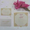 An open gold satin wedding invitation box displayed against a white background. The interior of the lid features a matching gold satin pocket holding a formal white invitation for "Mathew and Michelle" with gold-foil corner accents. The base of the box is lined with plush, gathered gold fabric. A stem of vibrant pink gladiolus flowers lies to the right, adding a pop of color to the scene.