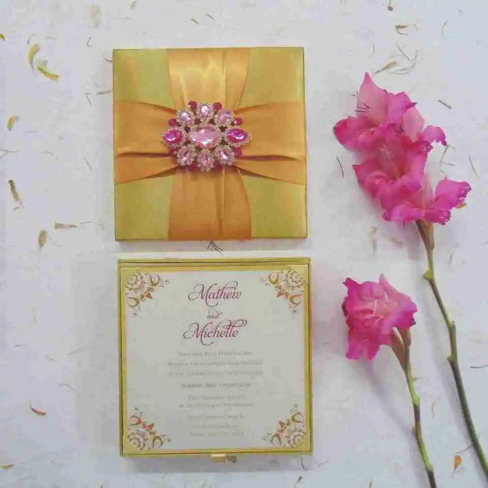 An open gold satin wedding invitation box displayed against a white background. The interior of the lid features a matching gold satin pocket holding a formal white invitation for "Mathew and Michelle" with gold-foil corner accents. The base of the box is lined with plush, gathered gold fabric. A stem of vibrant pink gladiolus flowers lies to the right, adding a pop of color to the scene.