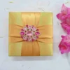 A top-down photograph of a closed square gold satin invitation box on a mottled white background with loose petals. The lid is wrapped with a wide, cross-shaped gold satin ribbon, cinched at the intersection by a large, ornate gold brooch heavily encrusted with sparkling clear and pink crystals in a starburst pattern. A cluster of magenta gladiolus flowers is visible to the right.