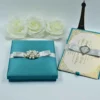 A top-down view of a closed, square Blue silk invitation box. The lid is elegantly decorated with a wide, matching silver satin ribbon wrapped horizontally across the center. At the midpoint of the ribbon, a large, ornate silver brooch is featured, intricately encrusted with shimmering clear crystals. The box is set against a clean, plain white background.