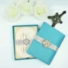 A top-down view of a closed, square Blue silk invitation box. The lid is elegantly decorated with a wide, matching silver satin ribbon wrapped horizontally across the center. At the midpoint of the ribbon, a large, ornate silver brooch is featured, intricately encrusted with shimmering clear crystals. The box is set against a clean, plain white background.