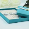 An open, square blue silk invitation box shown from a high angle. The interior lid features a matching horizontal silk pocket holding a white card with the name "Mathew and Michelle" in elegant gold script. The base of the box contains a formal wedding invitation card with gold-foil floral accents in the corners and matching gold lettering. The box is set against a clean white background.