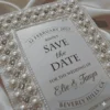 An ivory silk save the date card embroidered with faux pearls and rhinestones, with details printed with black foil on a pearl shimmery cardstock.