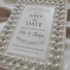 An ivory silk save the date card embroidered with faux pearls and rhinestones, with details printed with black foil on a pearl shimmery cardstock.